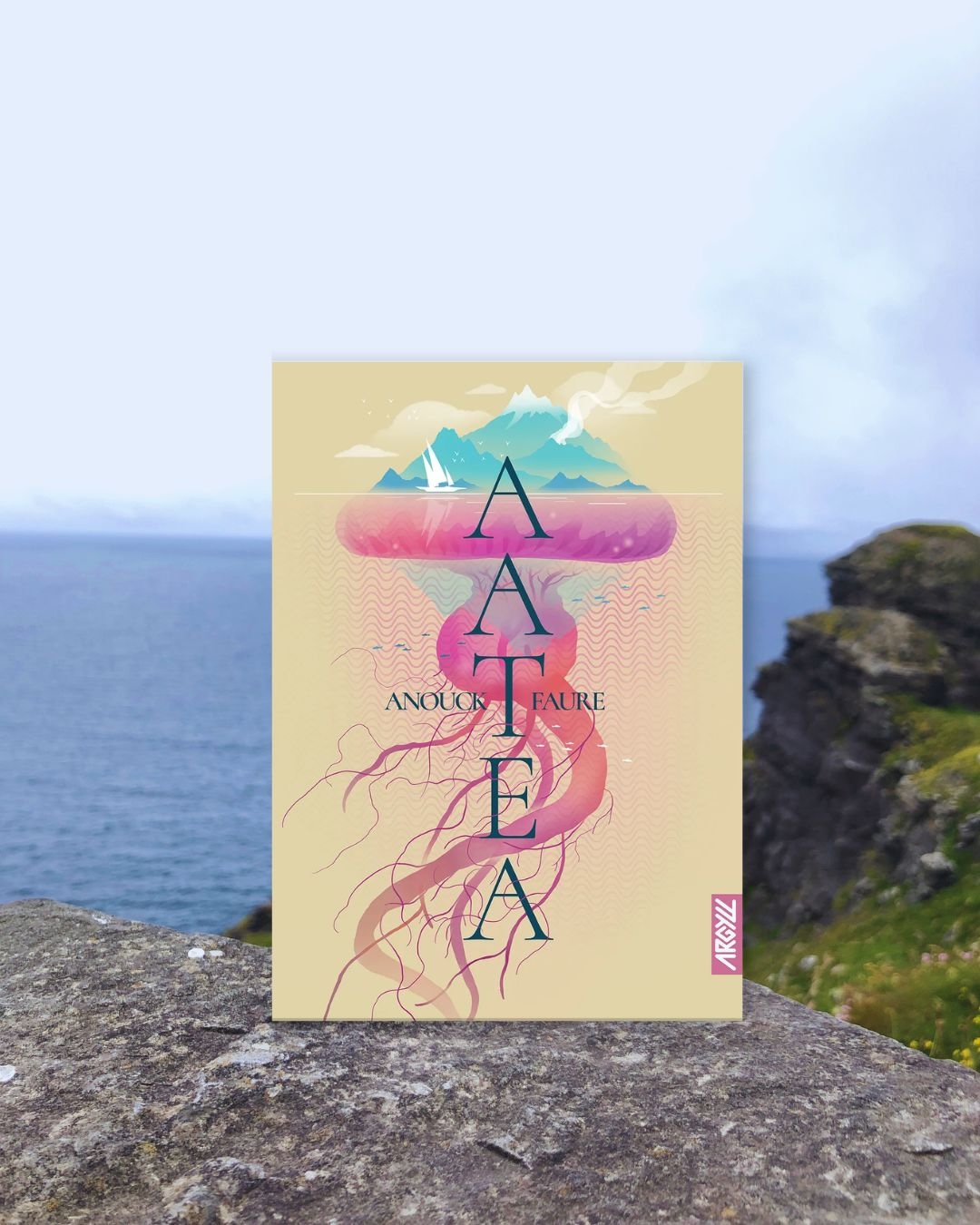 Livre posé sur un rocher devant la mer. La couverture d'Aatea montre une ile ressemblant à une méduse et un monocoque naviguant dans ses eaux.