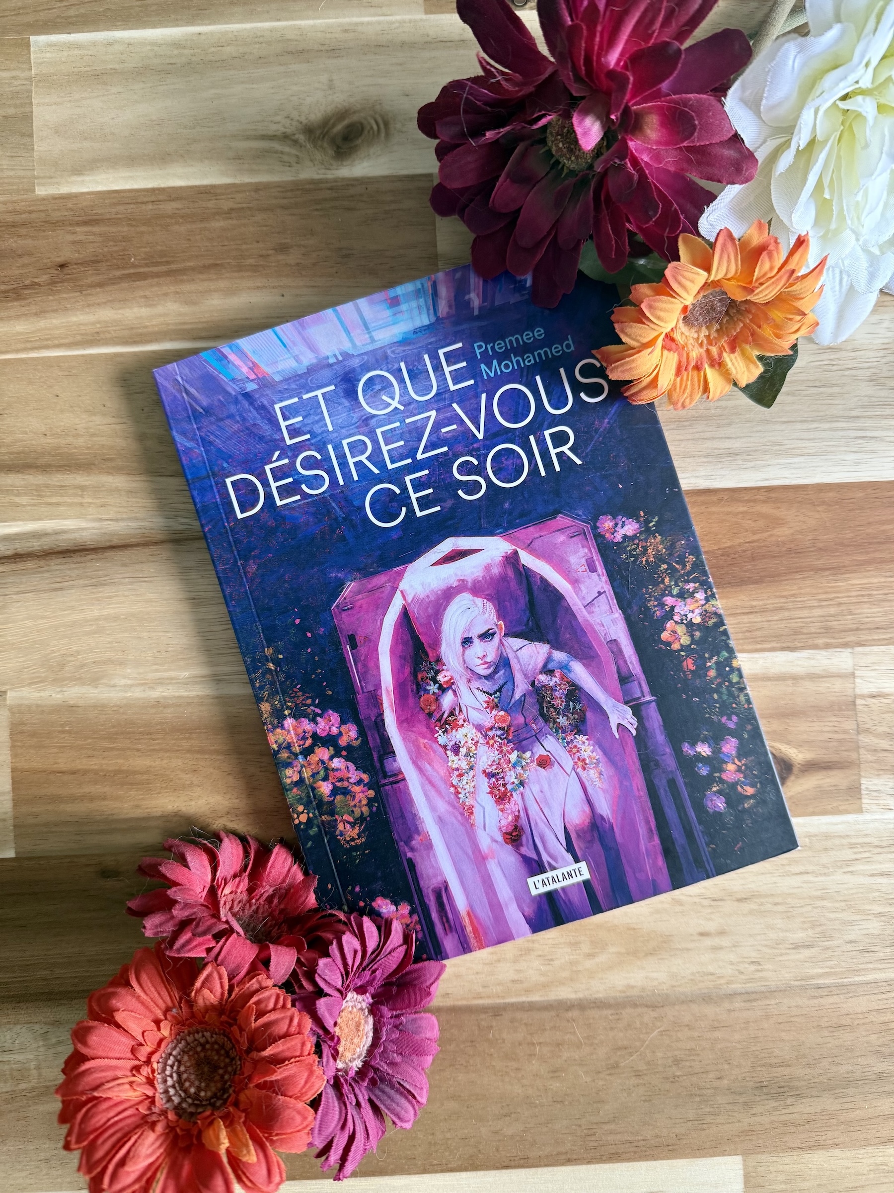 Livre posé avec des fleurs sur les côtés. La couverture de Et que désirez-vous ce soir montre une femme se relevant d’un cercueil entouré de fleurs. Son corps a les cicatrices d’une autopsie qui craquent et d’où s’écoulent des fleurs. 