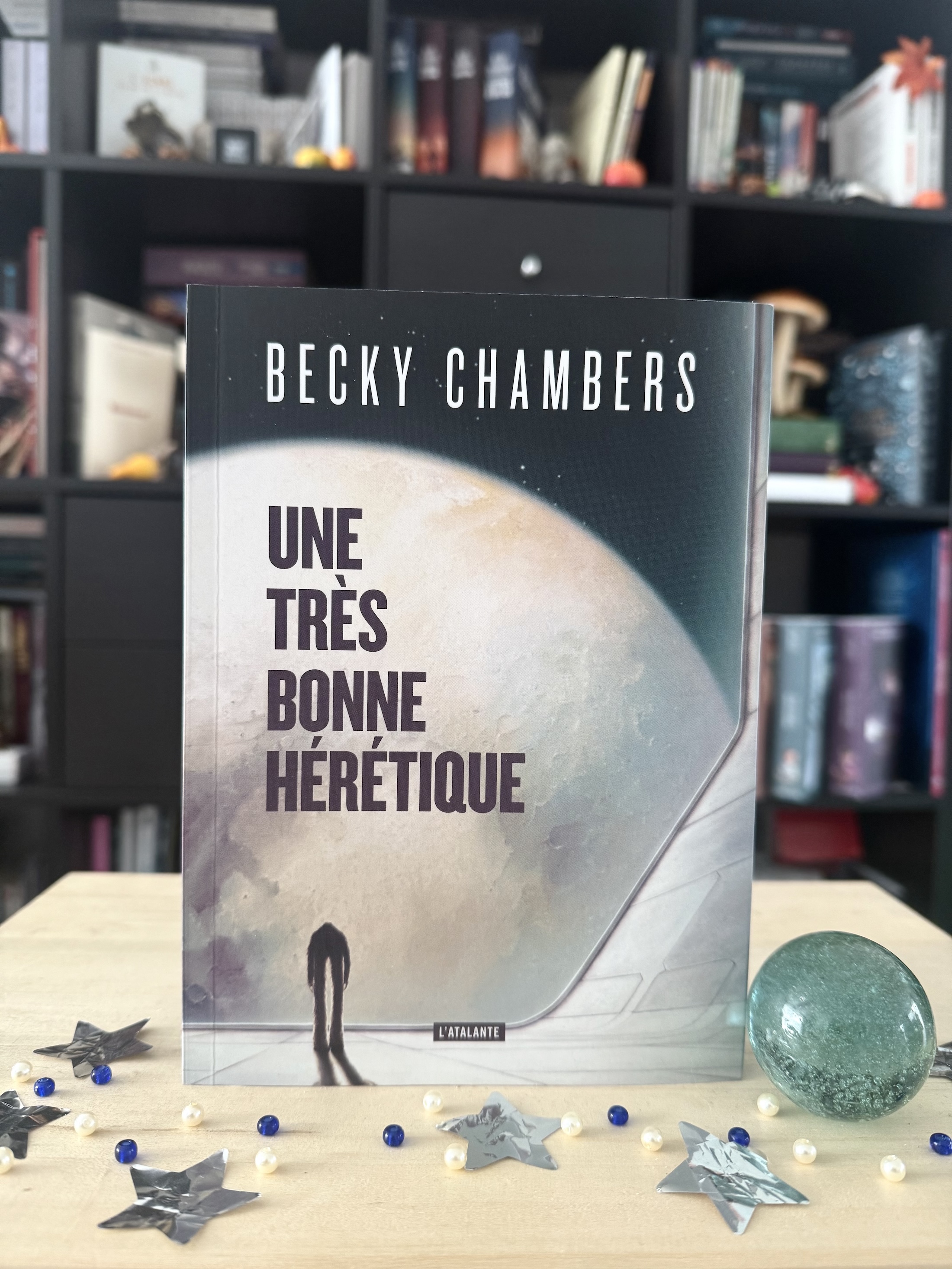 Livre posé devant une bibliothèque avec des étoiles et des perles blanches et bleues ainsi qu’un calot qui représente une planète. La couverture d’Une très bonne hérétique montre une créature bipède sur une surface de vaisseau/station spatiale avec en face une planète.