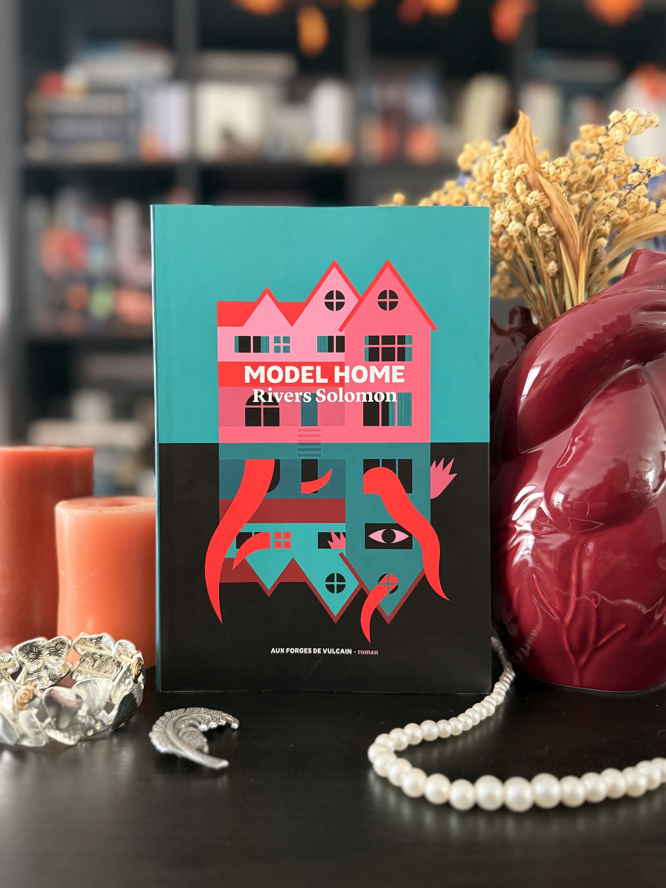 Livre posé devant une bibliothèque avec des bijoux, des bougies, un vase en forme de coeur anatomique avec des fleurs séchées dedans. La couverture du livre montre une belle maison avec en miroir, la même bâtisse à l’envers d’où s’échappent des tentacules et bras géants et un oeil à une fenêtre. 