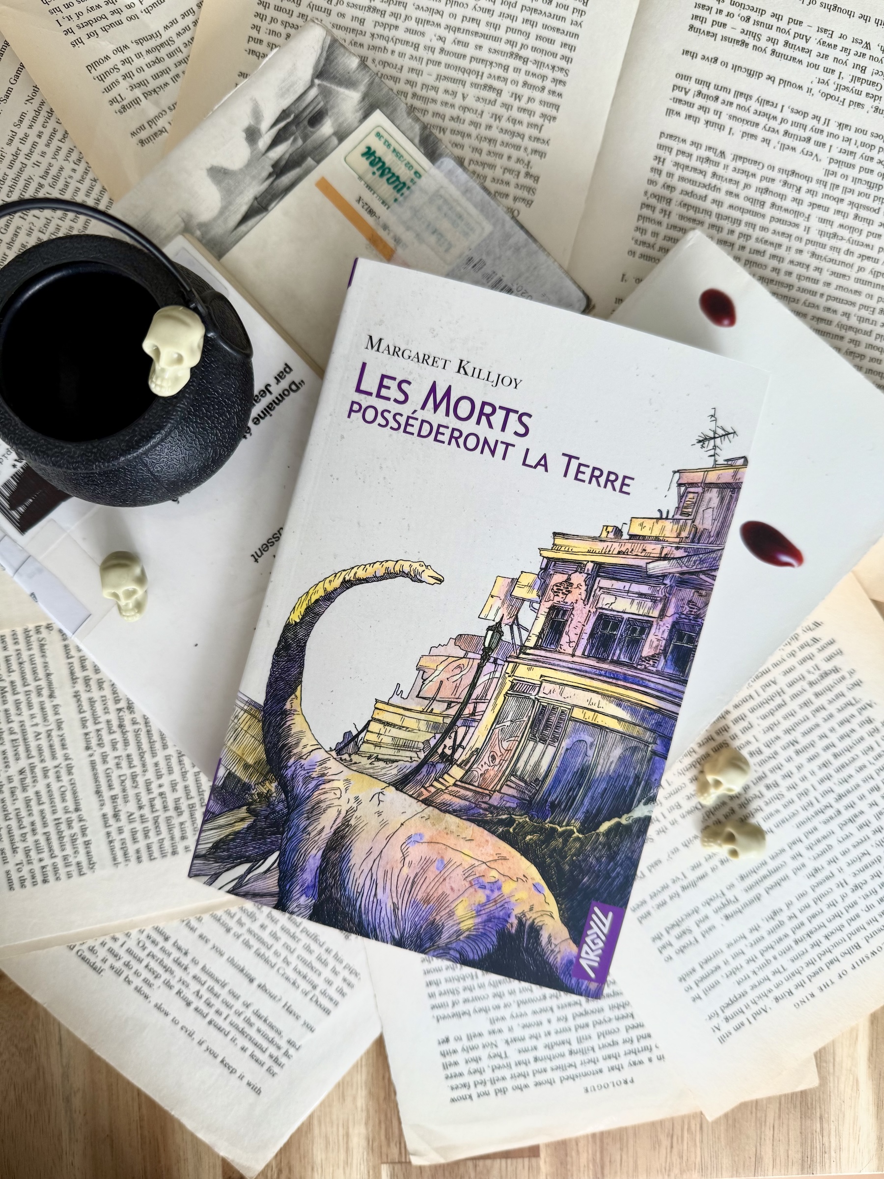 Livre posé sur des livres de bibliothèque et des pages volantes de livres, aux côtés d'un mini-chaudron et de petites têtes de morts. La couverture de Les morts posséderont la terre montre une dinosaure en béton devant des bâtiments anciens usés.