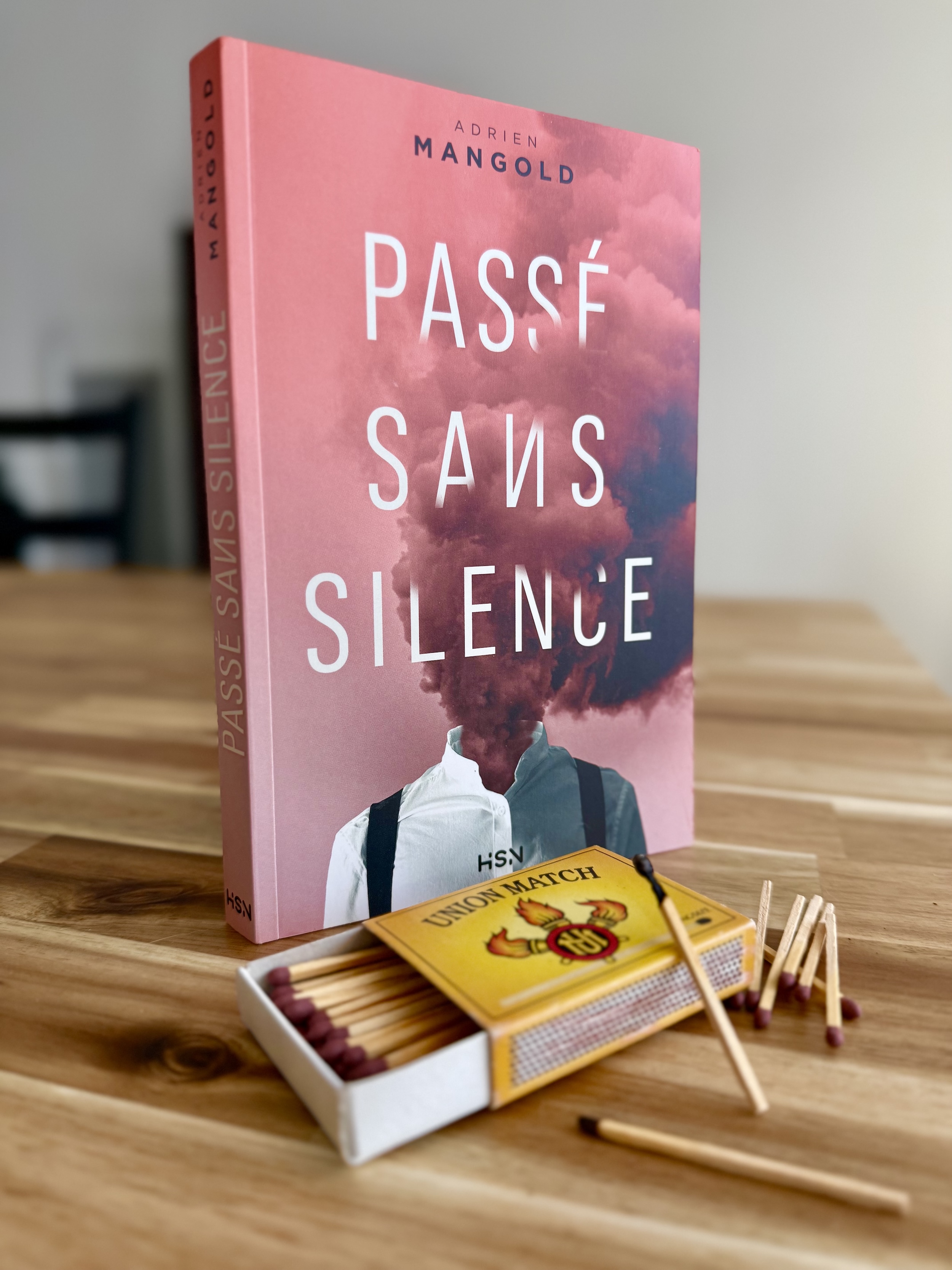 Livre posé sur une table avec devant une boite d'allumettes dont une déjà brûlée. La couverture du livre montre une silhouette en chemise blanche dont la tête a été remplacée par une fumée orangée-rouge 