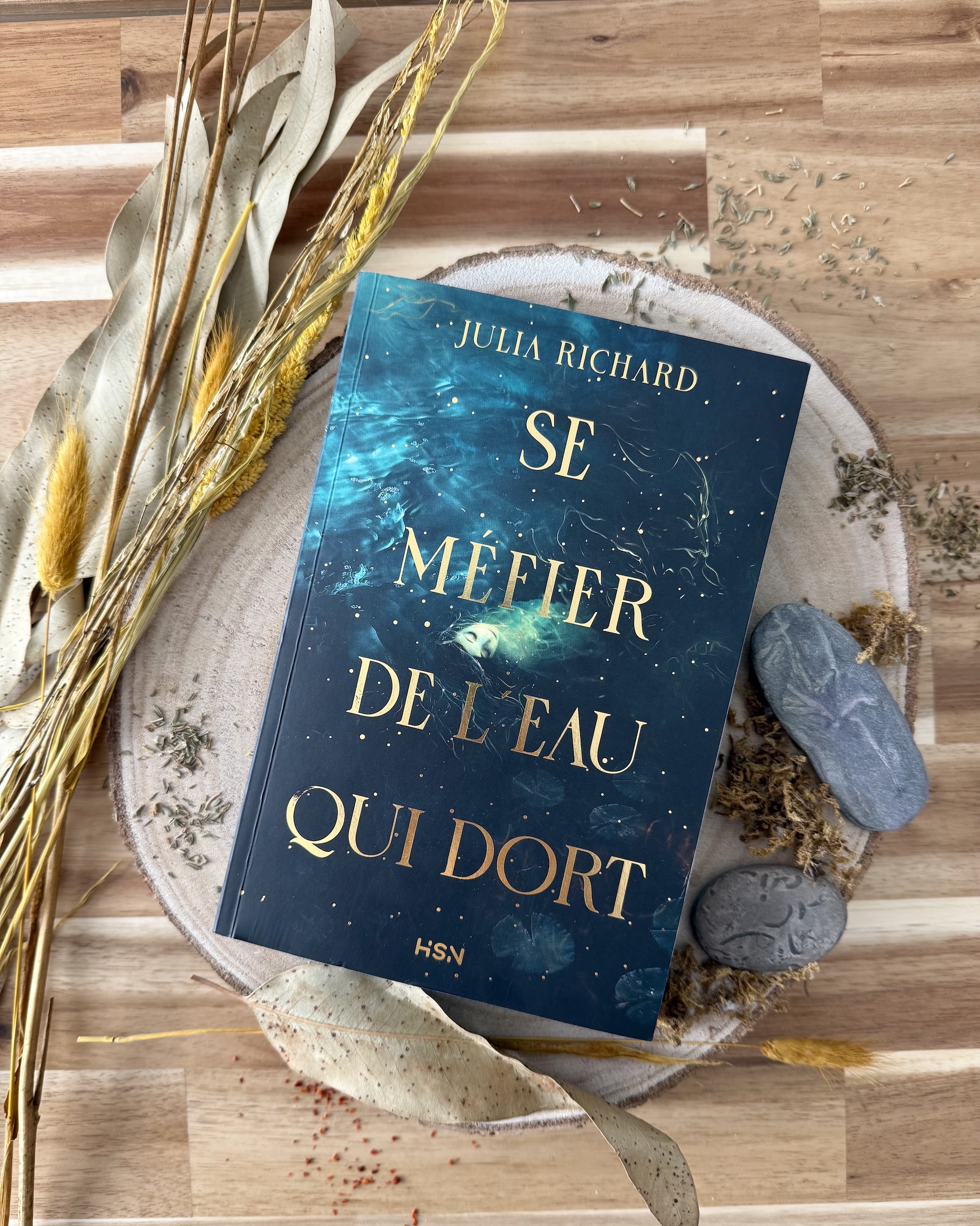 Livre posé sur un rondin de bois entouré de pierres, de mousse, d'épices et herbes séchées et de fleurs séchées. La couverture de Se méfier de l’eau qui dort montre un personnage aux traits féminins, allongé dans une rivière un peu agitée. Des particules dorées parsèment la page et accompagnent la dorure brillante du titre et du nom de l’autrice.